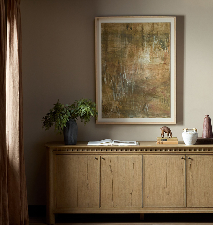 A large framed abstract painting with earthy brown, gold, and beige tones hangs on a taupe wall above a light wood sideboard. The room includes a dark blue vase with greenery, an open book, a small elephant figurine, books, and a white pitcher.