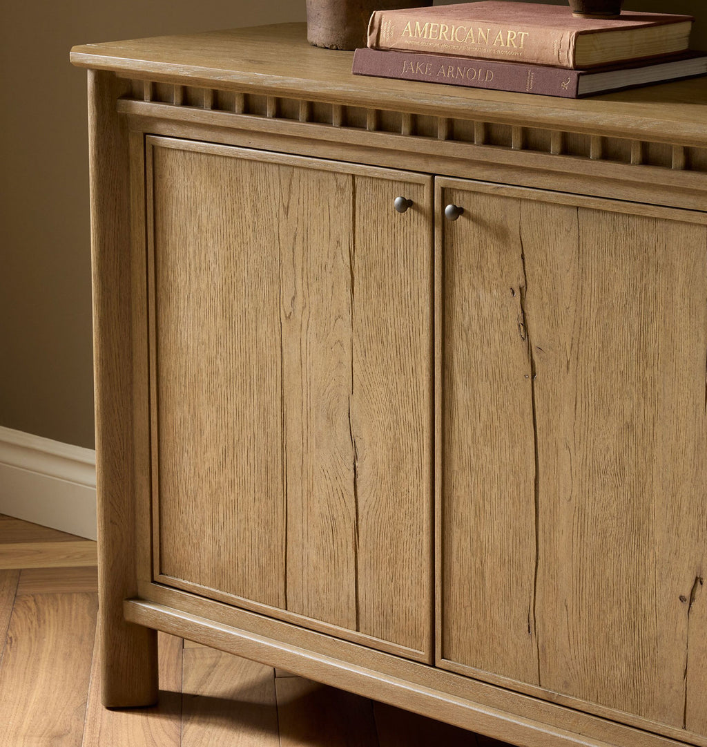 A close-up of a rustic light brown wooden cabinet with two doors and small round metal knobs, set against a warm brown wall and wood floor. Books are stacked on top.