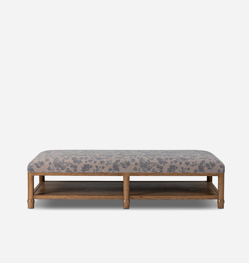 A rectangular upholstered bench with a medium brown wooden frame and lower shelf, featuring a gray and beige floral patterned fabric cushion, shown against a light gray background.