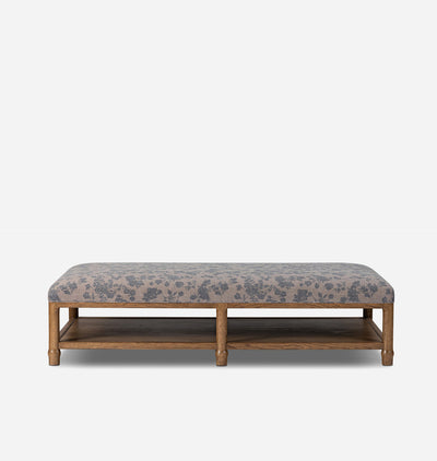 A rectangular upholstered bench with a medium brown wooden frame and lower shelf, featuring a gray and beige floral patterned fabric cushion, shown against a light gray background.