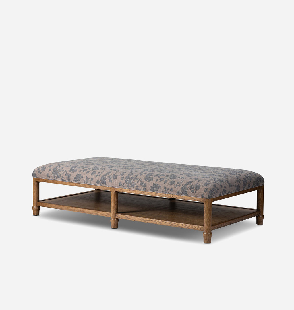 A rectangular upholstered bench with a medium brown wooden frame and lower shelf, featuring a gray and beige floral patterned fabric cushion, shown against a light gray background.