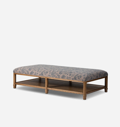 A rectangular upholstered bench with a medium brown wooden frame and lower shelf, featuring a gray and beige floral patterned fabric cushion, shown against a light gray background.