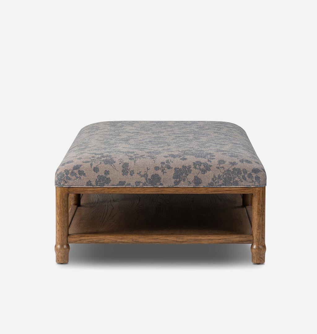 A rectangular upholstered bench with a medium brown wooden frame and lower shelf, featuring a gray and beige floral patterned fabric cushion, shown against a light gray background.