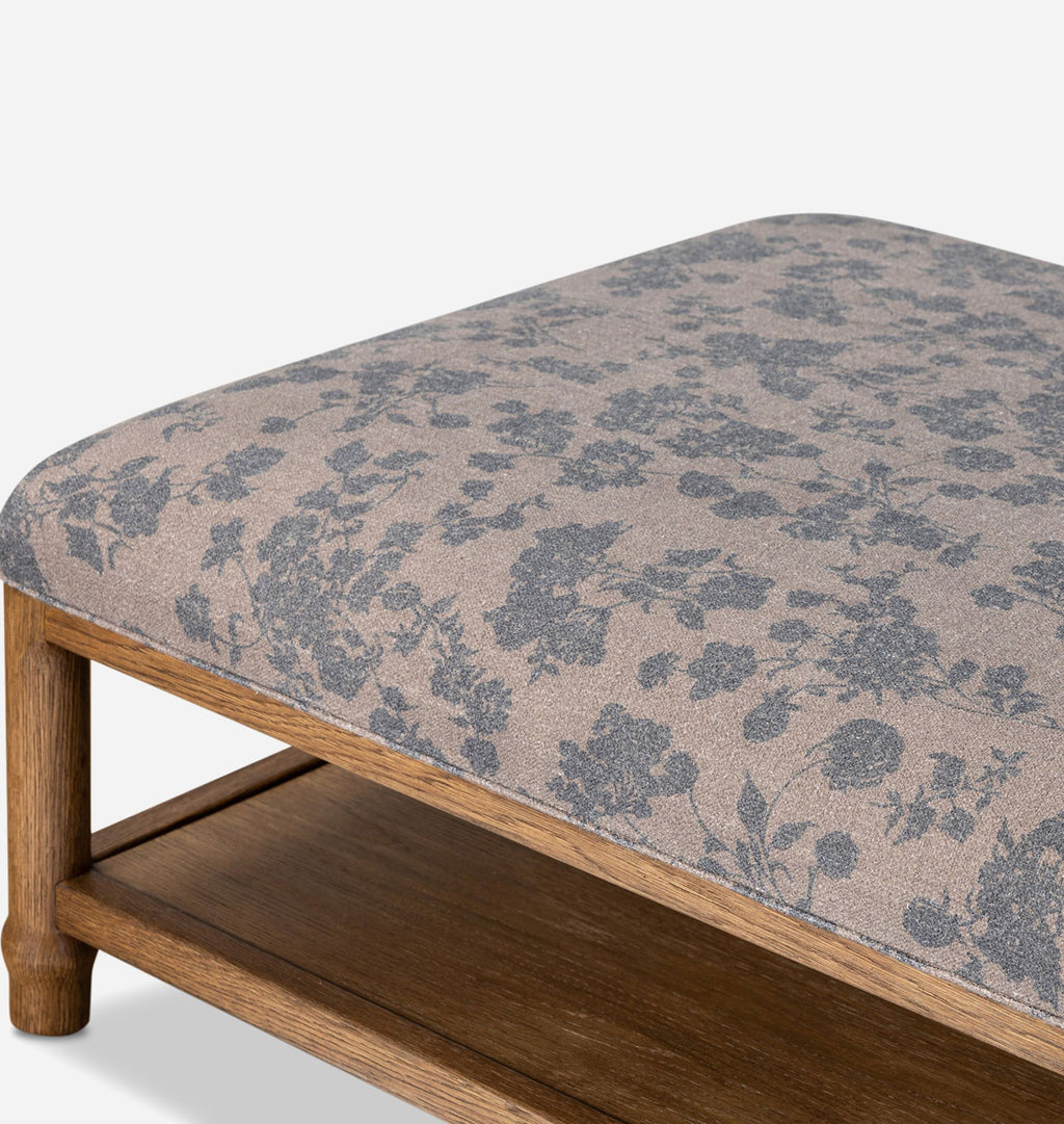 A close-up of a rectangular upholstered ottoman or bench cushion with a muted floral pattern in gray and taupe, mounted on a medium brown wooden frame with a lower shelf.