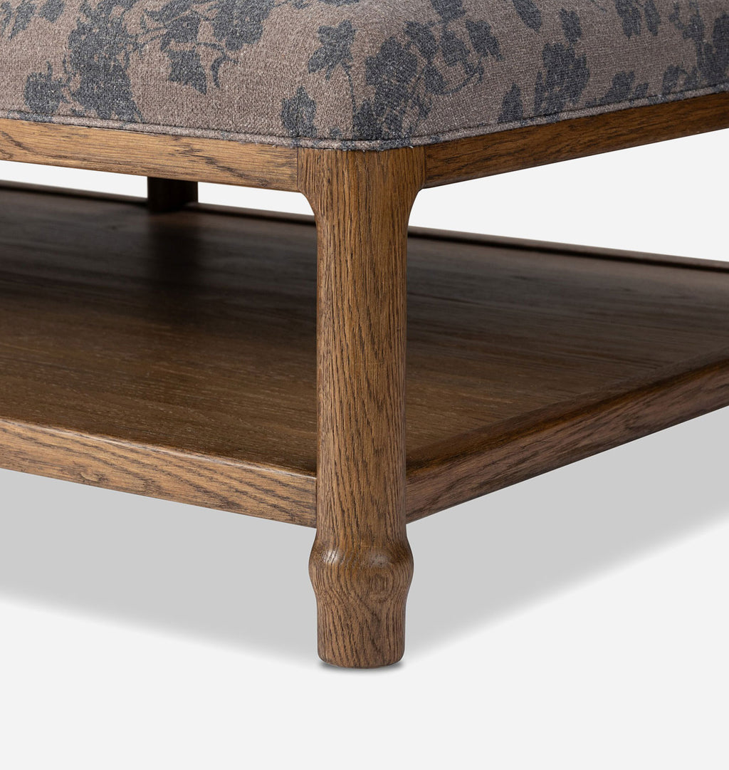 Close-up of a wooden ottoman or bench leg with a patterned upholstered seat cushion and a dark wood shelf, shown on a light gray studio background.