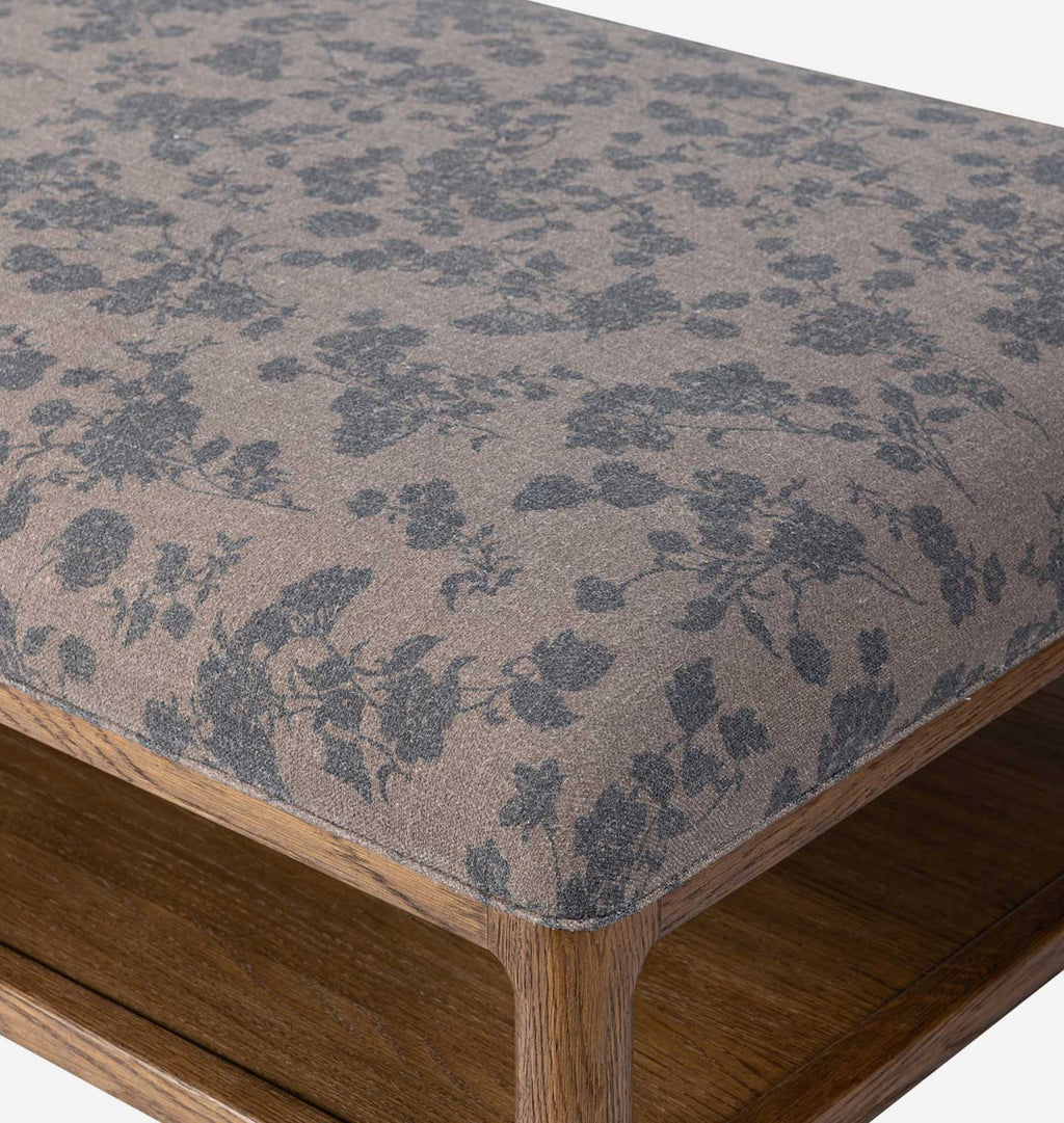 Close-up of an upholstered wooden ottoman or bench seat with a taupe fabric top featuring a dark gray floral toile pattern, showing the wooden frame and leg.