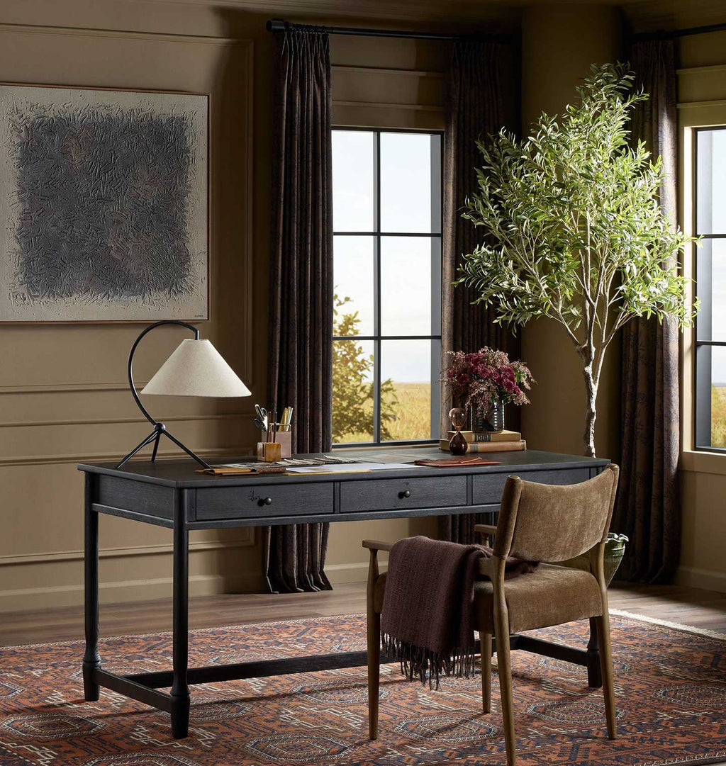 A dark gray writing desk in a styled home office setting, paired with a brown upholstered wooden chair draped with a dark throw blanket. The scene includes a black metal desk lamp, a patterned area rug, a potted olive tree, and large windows with dark curtains.