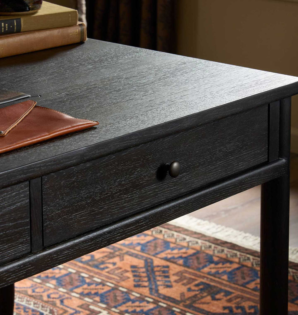 A close-up of a dark wood writing desk with a single drawer and a round metal knob, shown in a home office setting with books, a leather notebook, and a patterned rug.