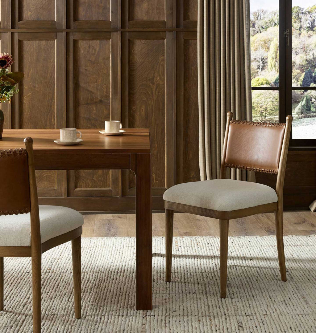 A dining room scene showing two wooden chairs with cream upholstered seats and brown leather backrests, placed around a matching wooden table in a warm interior with wood paneling and a window.