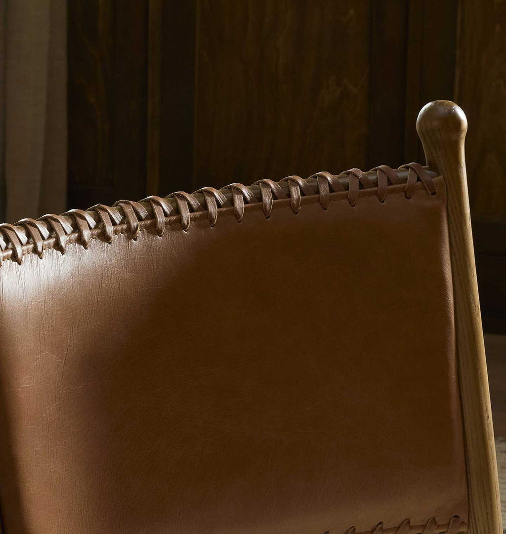 Close-up of a brown leather chair back with whipstitching and a light wood frame, set against a dark wood-paneled background.