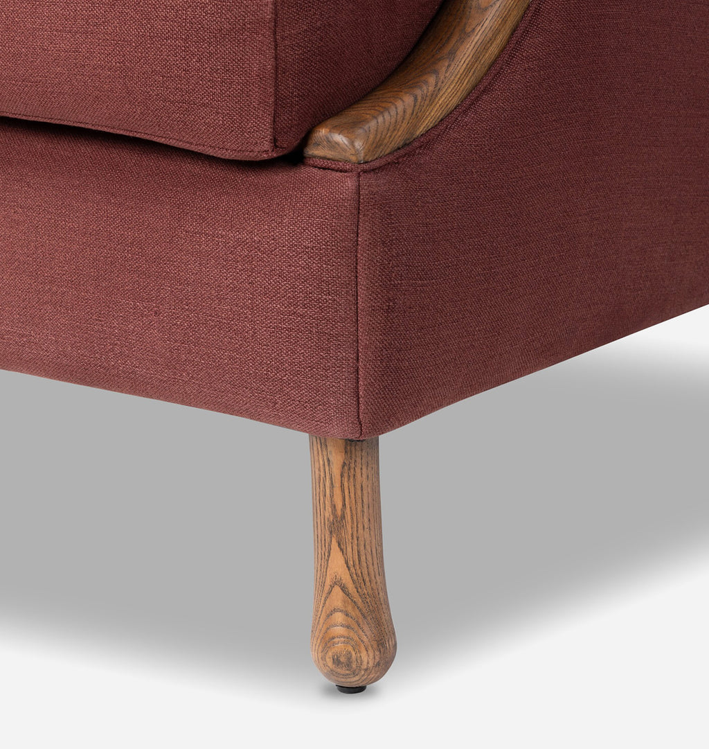 Close-up of a upholstered armchair corner showing rust-red fabric, a curved wooden arm, and a turned wood leg against a light gray studio background.