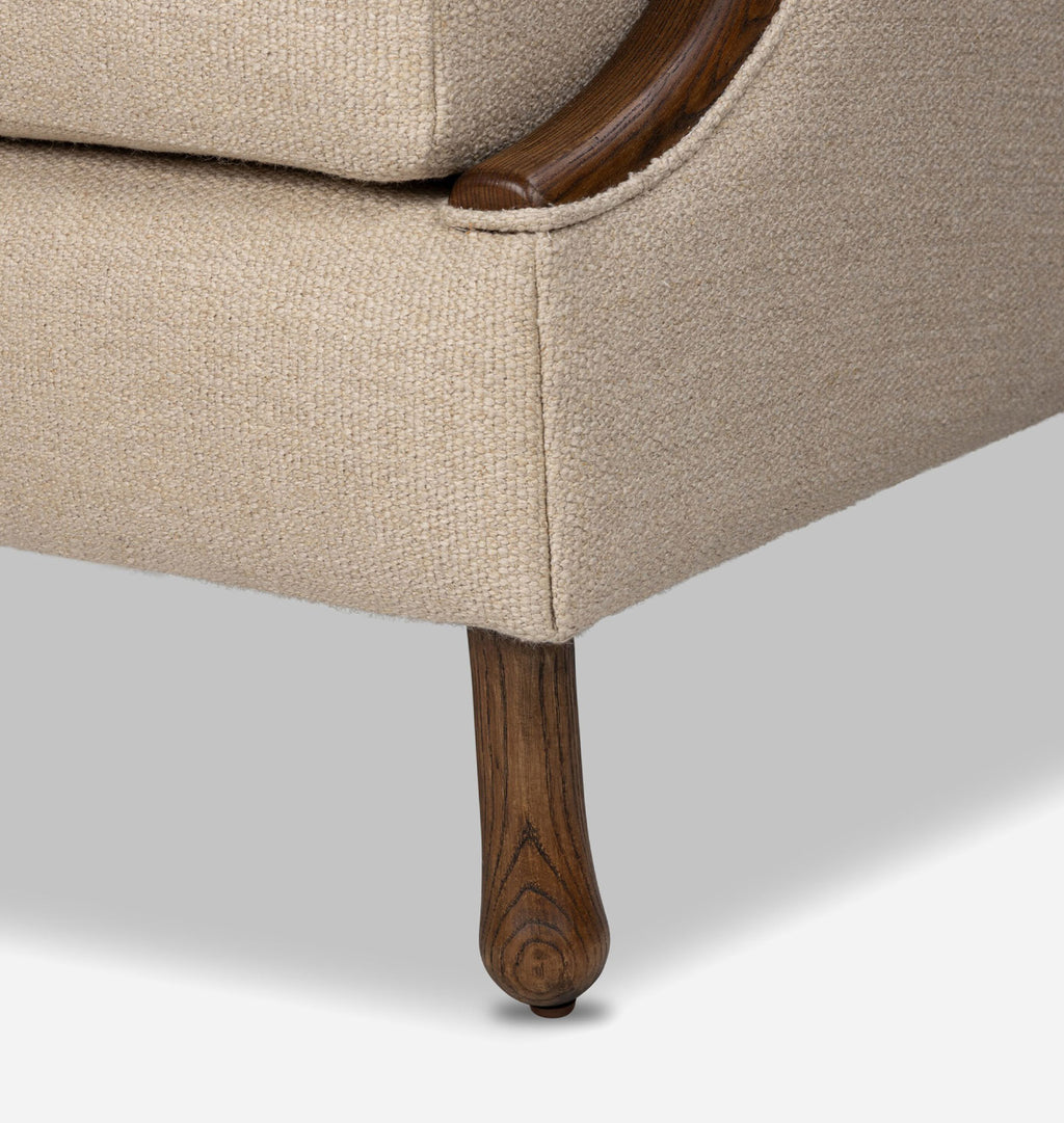 Close-up detail of an upholstered chair or sofa corner, showing beige woven fabric, a dark wood armrest, and a turned wooden leg on a light gray studio background.