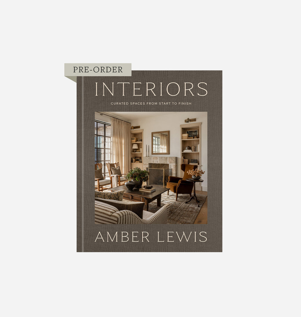 The front cover of the book 'Interiors: Curated Spaces from Start to Finish' by Amber Lewis, shown on a light gray background. The cover is a muted taupe-brown with a central photograph of a cozy living room and a beige 'PRE-ORDER' tab on the upper left.
