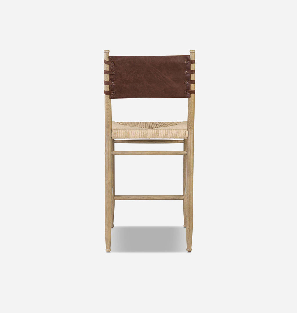 A modern wooden barstool with a light natural wood frame, a woven natural fiber seat, and a dark brown leather backrest strap, shown from the back against a light gray background.