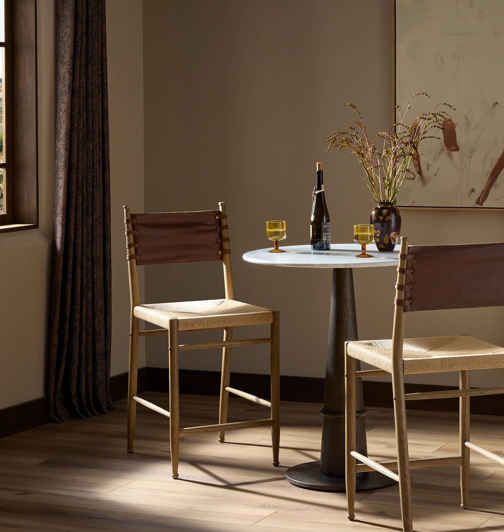 Two wooden bar stools with woven beige seats and brown leather backs are arranged around a small round table in a warmly lit room with a window, curtains, and a vase of branches.