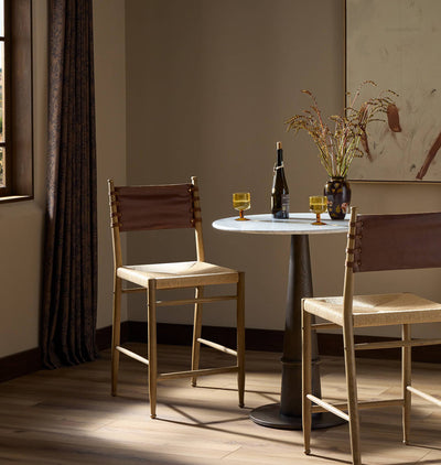 Two wooden bar stools with woven beige seats and brown leather backs are arranged around a small round table in a warmly lit room with a window, curtains, and a vase of branches.