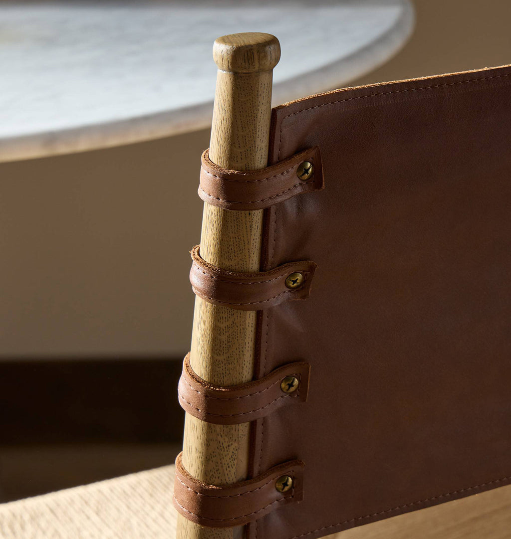 Close-up of a wooden chair back with a brown leather sling strap secured by four leather straps and brass grommets, attached to a light wood post.
