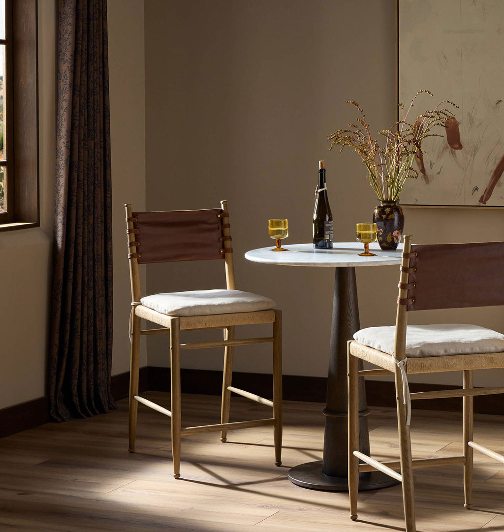 Two modern bar stools with light wood frames, brown leather backrests, and cream cushions are arranged around a small round table in a warmly lit room with a window, curtains, and a vase of branches.