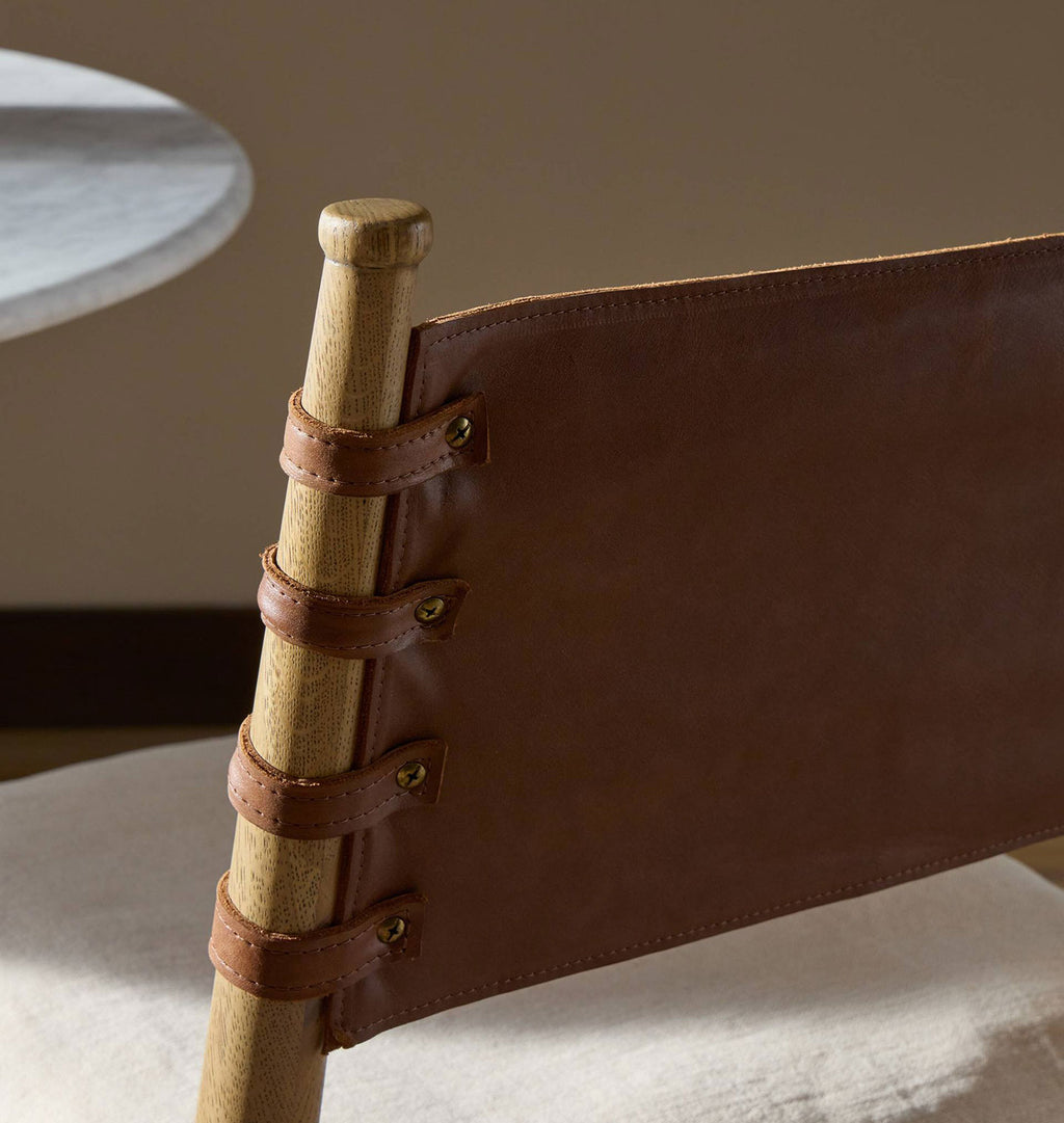 Close-up of a wooden chair back with a brown leather sling strap secured by four leather loops and brass grommets, set in a warm neutral interior.