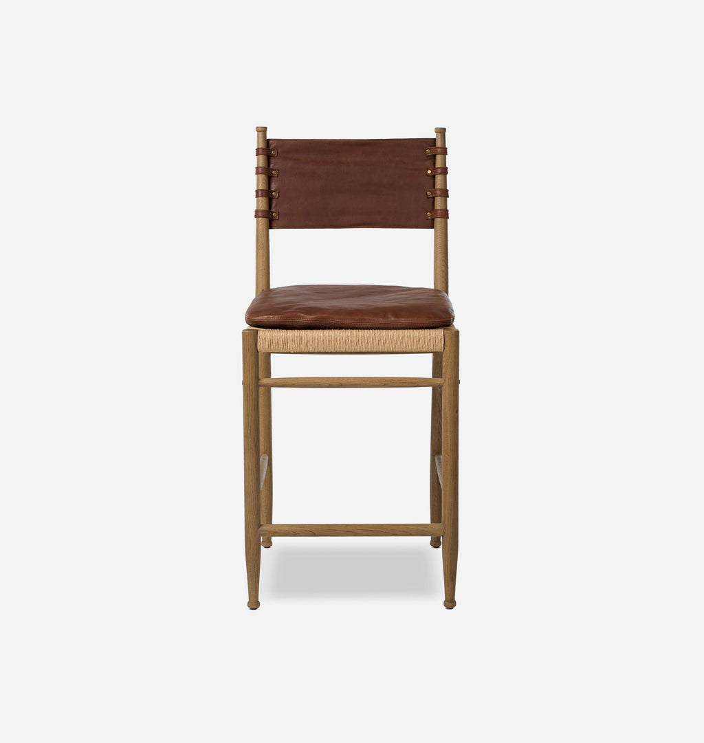 A mid-century modern counter stool featuring a light natural wood frame, a brown leather sling backrest, and a brown leather seat cushion over a woven cane base, shown against a plain light gray background.