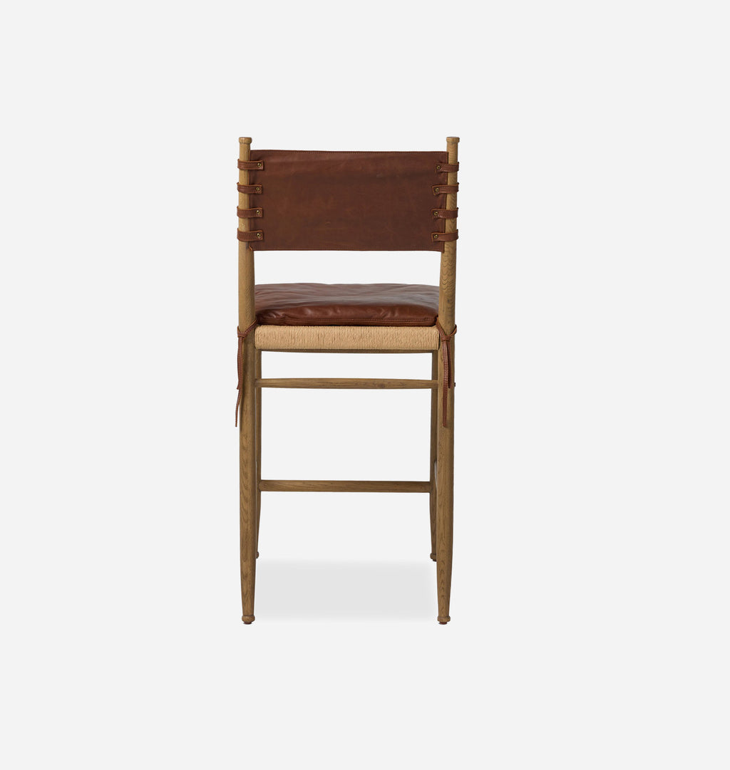 A wooden bar stool with a brown leather seat cushion, a woven cane seat, and a backrest featuring vertical brown leather straps tied to the wooden frame, shown against a light gray background.