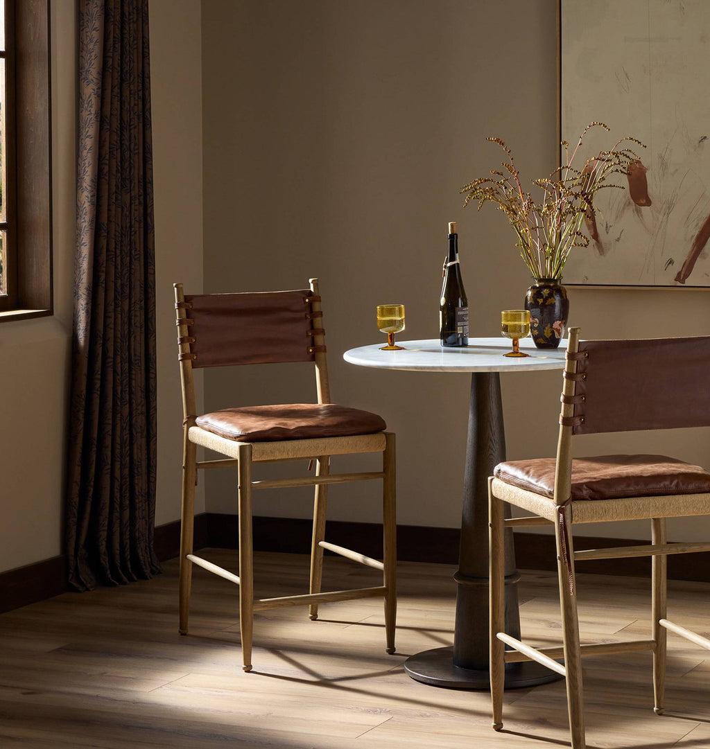 Two wooden bar stools with brown leather seats and backs are arranged around a small round table in a warmly lit room with curtains, a vase of branches, and a bottle on a marble-top table.