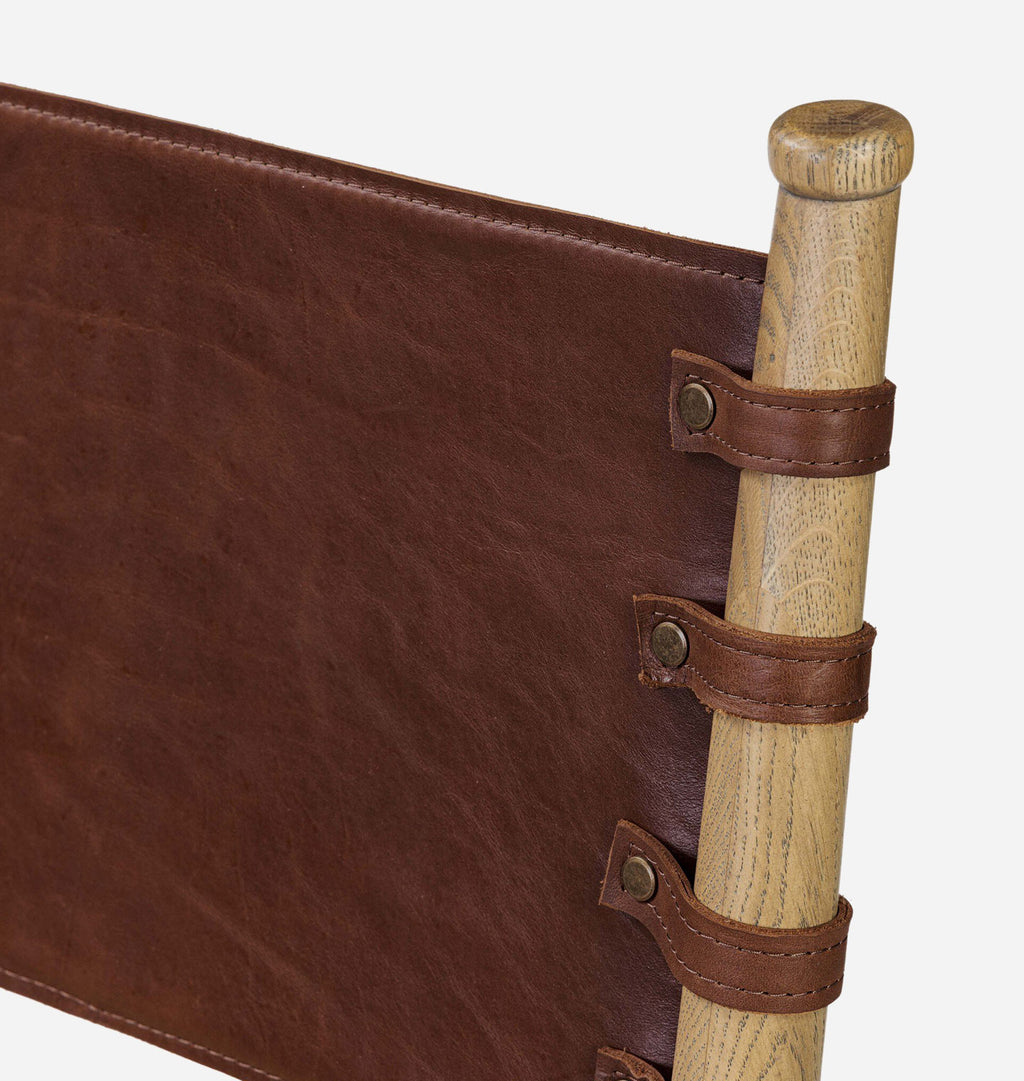 Close-up of a brown leather sling seat or hammock strap attached to a light natural wood pole with metal rivets and leather loops, shown on a plain white background.