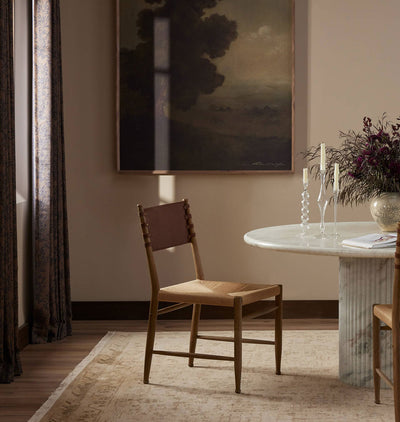 A wooden dining chair with a woven seat and brown leather backrest, placed in a styled dining room next to a marble-topped table with candles and a framed landscape painting on the wall.