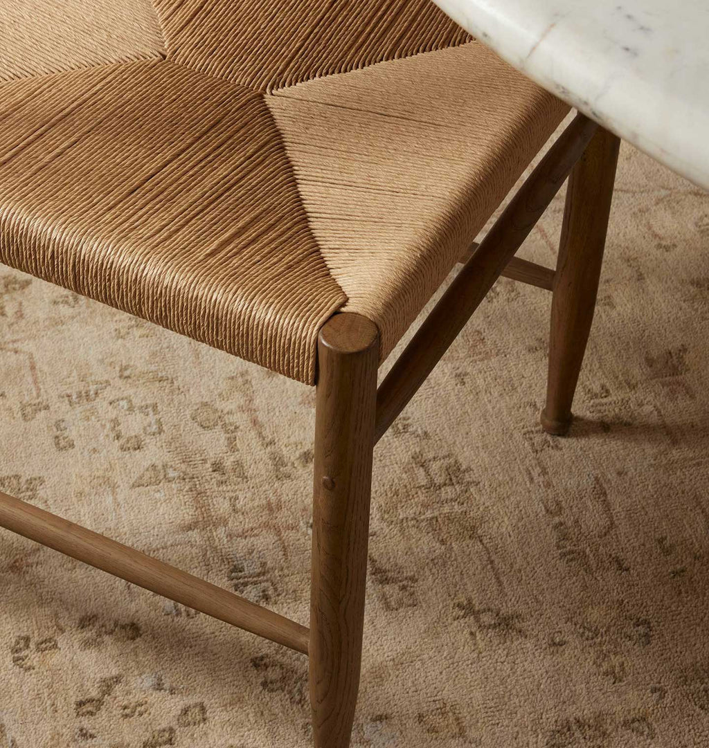 Close-up of a mid-century modern chair with a woven natural-fiber seat and warm brown wooden frame, shown on a patterned beige rug with a marble tabletop edge visible in the corner.