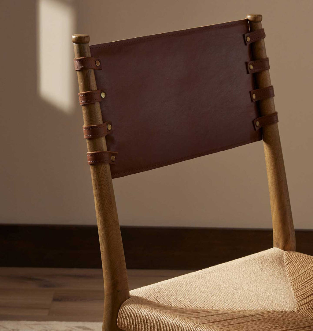 A close-up of a wooden chair with a dark brown leather backrest strap and a woven beige seat, set against a neutral indoor wall and wood floor.