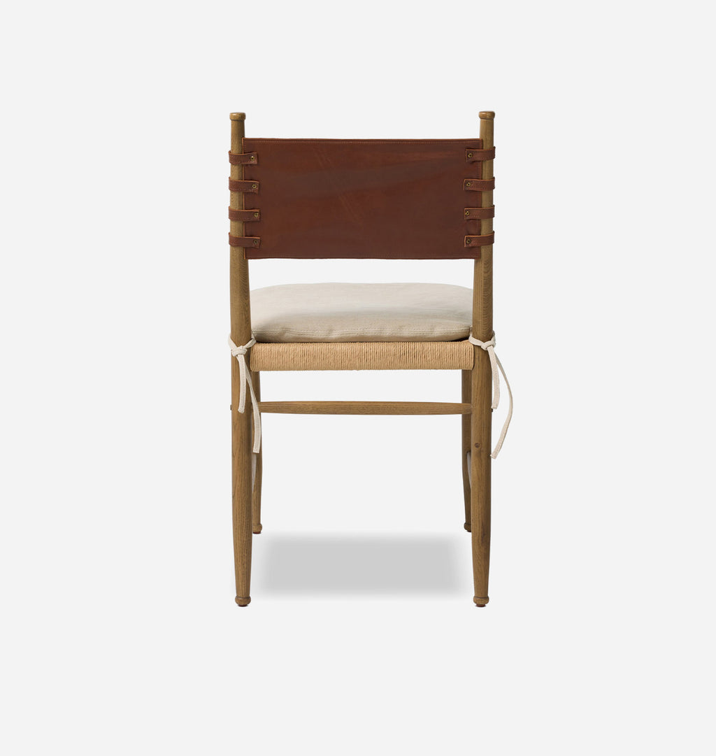 A wooden dining chair with a brown leather back strap, a beige woven seat, and a cream cushion, shown from the front against a light gray background.