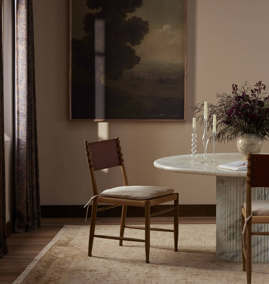 A styled dining room scene featuring a wooden dining chair with a brown leather backrest and a light beige upholstered seat, positioned next to a marble-topped table with candles and a floral arrangement.