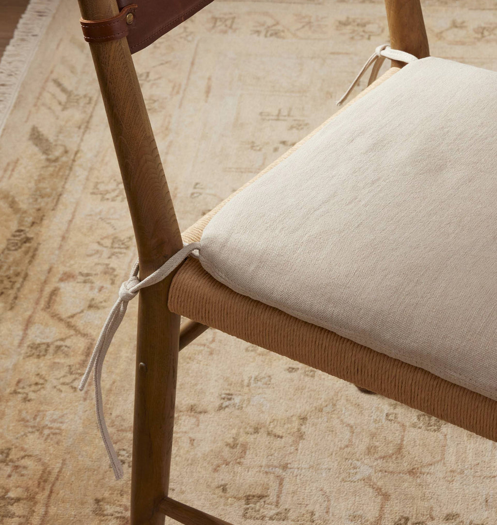 A close-up of a cream-colored fabric chair cushion tied to a wooden chair frame, set on a patterned beige rug.