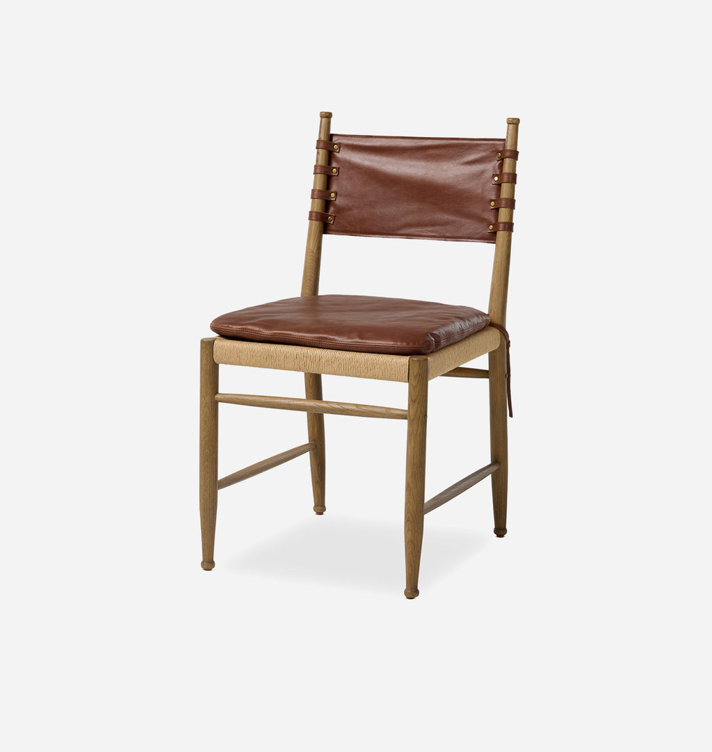 A wooden dining chair with a brown leather seat cushion and backrest, featuring vertical leather straps on the sides, set against a plain light gray background.