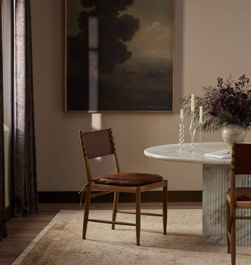 A styled dining room scene featuring a wooden dining chair with a brown leather seat and backrest, positioned next to a marble-topped table with candles and a floral arrangement.