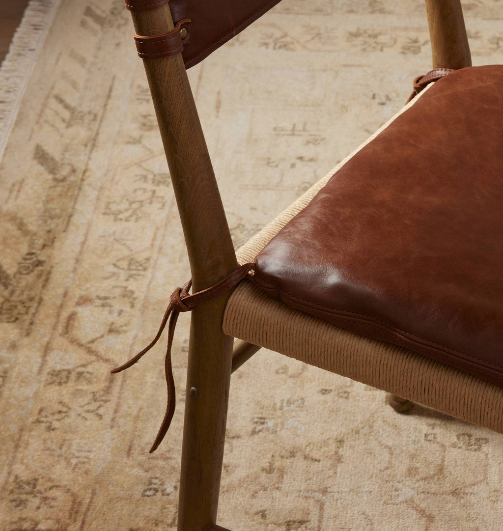 A close-up of a wooden chair with a brown leather seat cushion tied with leather straps, set on a patterned beige rug.