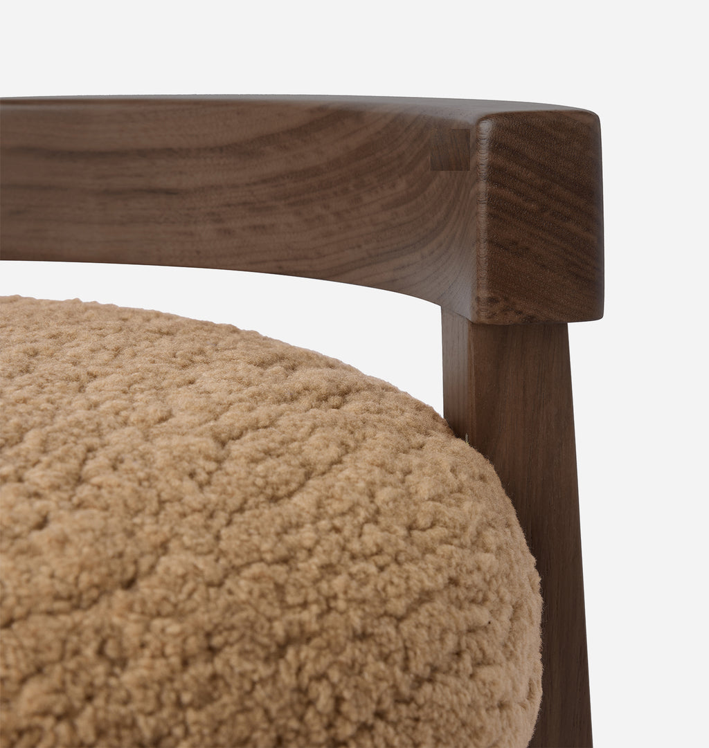 Close-up detail of a wooden chair arm and leg with a textured beige shearling upholstered seat against a light gray background.
