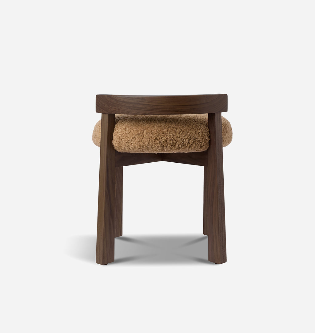 A modern wooden stool with a dark brown walnut frame and a round, textured tan shearling seat cushion, shown from the front on a light gray background.