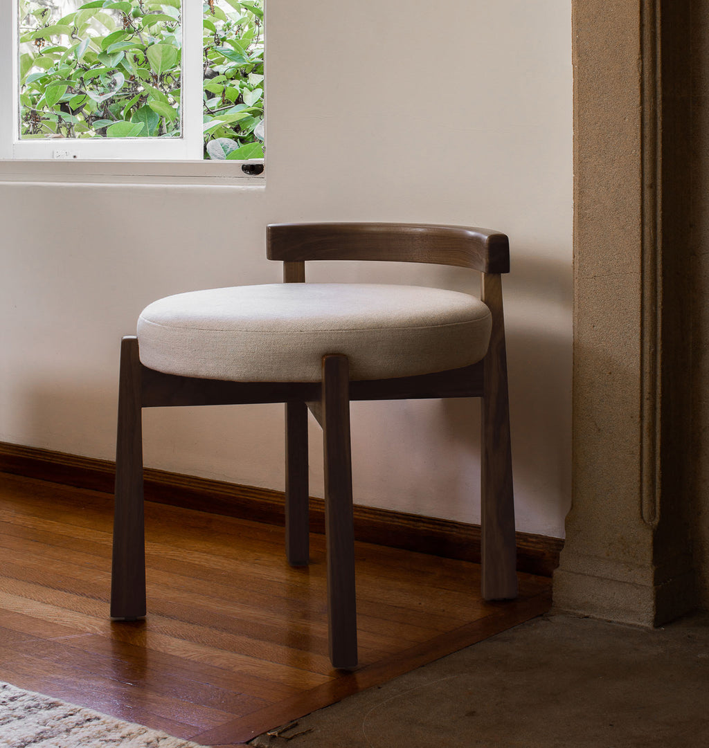 A modern wooden dining chair with a curved backrest and a round off-white upholstered seat cushion, positioned in a room corner with a wooden floor, beige wall, and window.