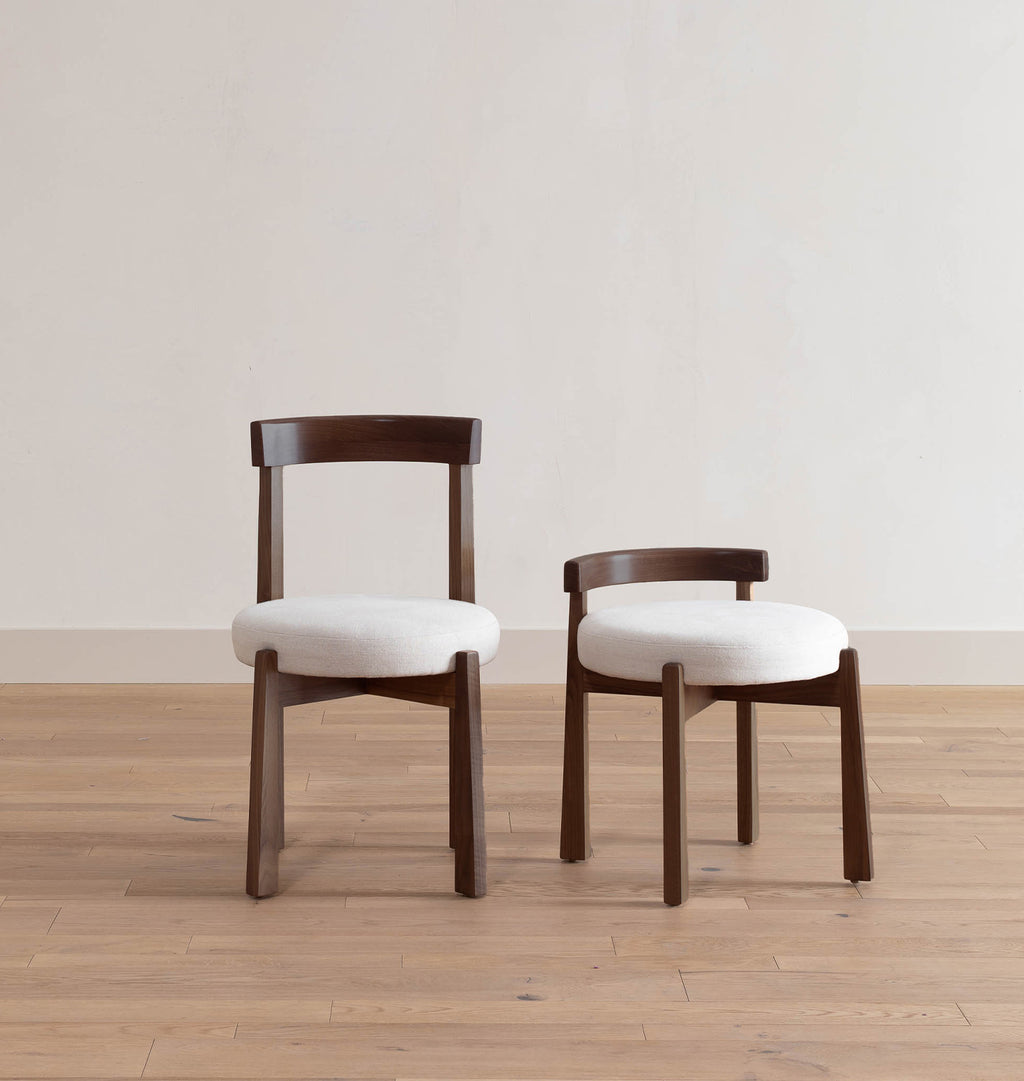 Two wooden dining chairs with white upholstered seats, shown in a minimalist room with light wood floors and a plain off-white wall.