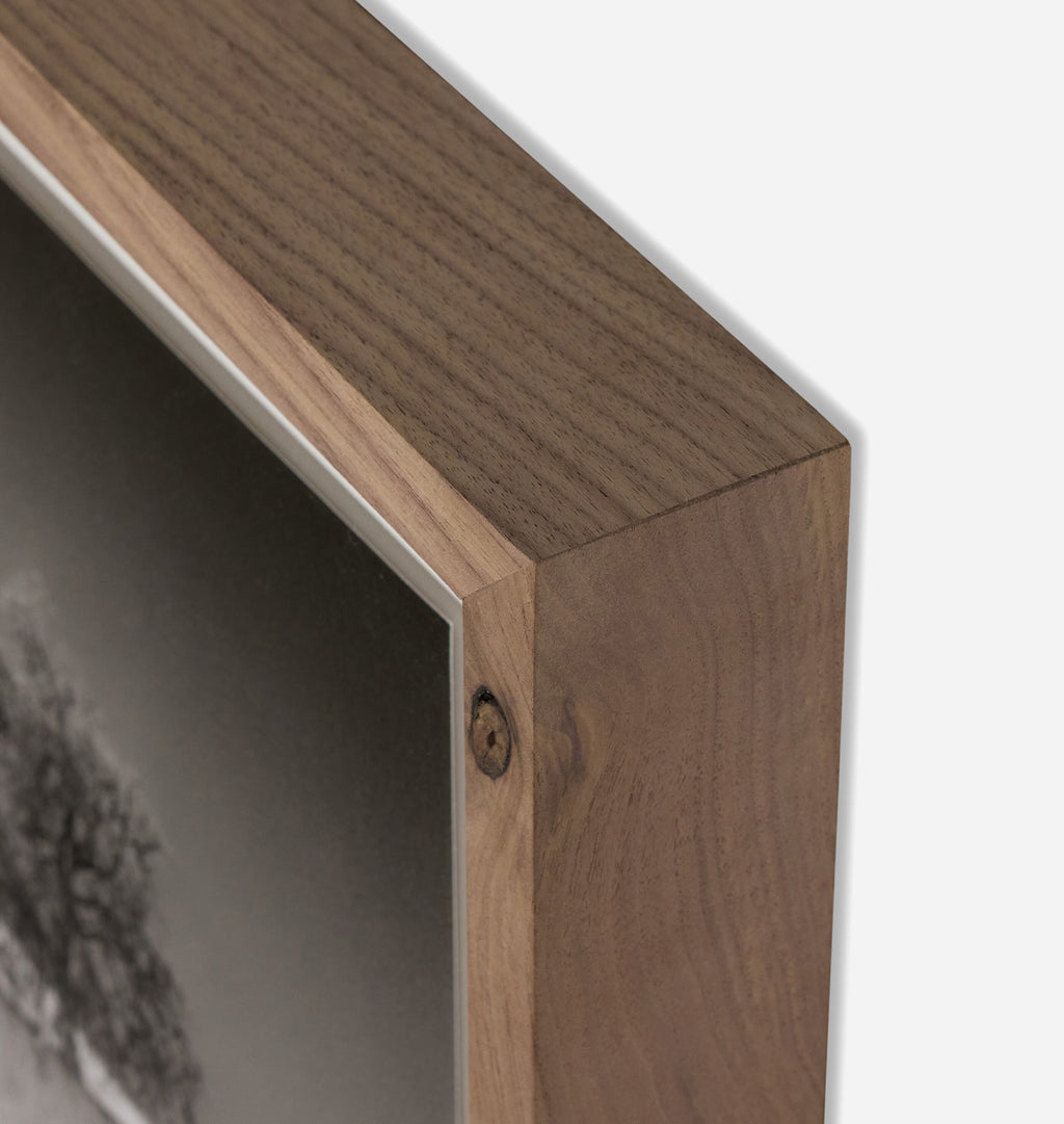 Close-up of a framed wall art piece showing the wooden frame corner with a visible wood grain and a mounted print inside, against a light neutral background.