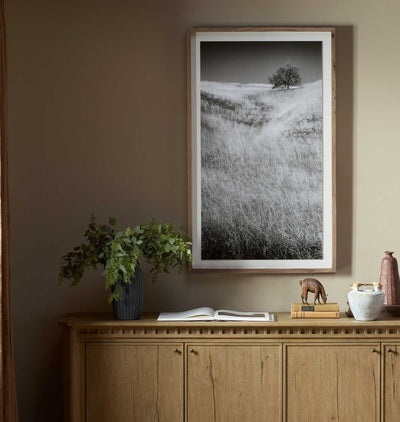 A framed black and white landscape photograph of a grassy hill with a lone tree, displayed on a warm brown wall above a wooden console table with decorative items.