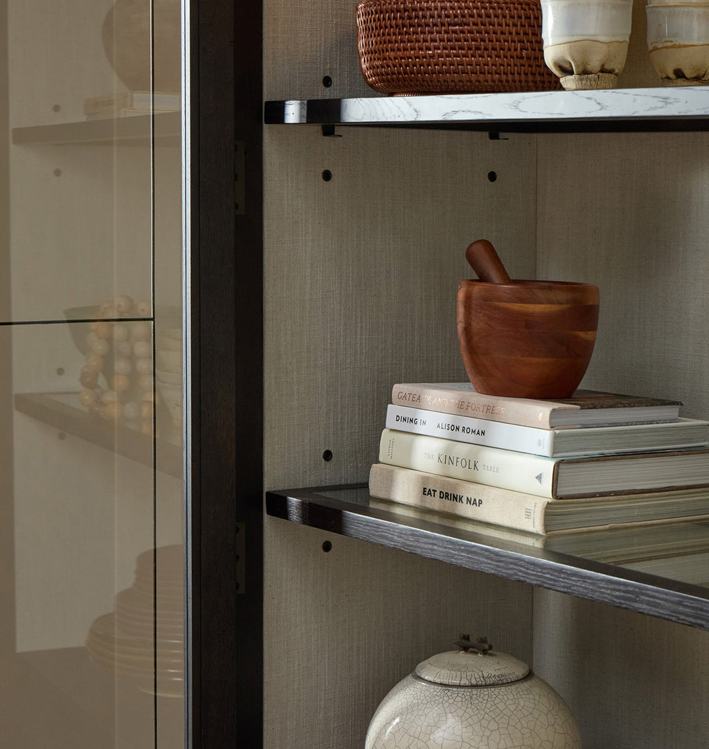 A close-up of a modern interior shelving unit with floating shelves, a glass cabinet door, and decorative objects including books, a wooden mortar and pestle, ceramic vessels, and woven baskets.