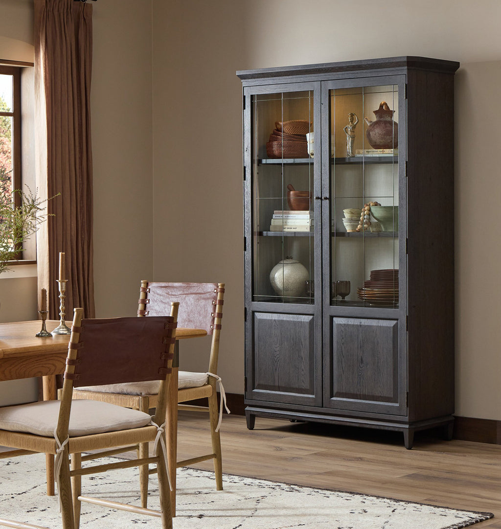 A tall dark gray wood china cabinet with glass doors stands in a dining room next to a wooden table and chairs.