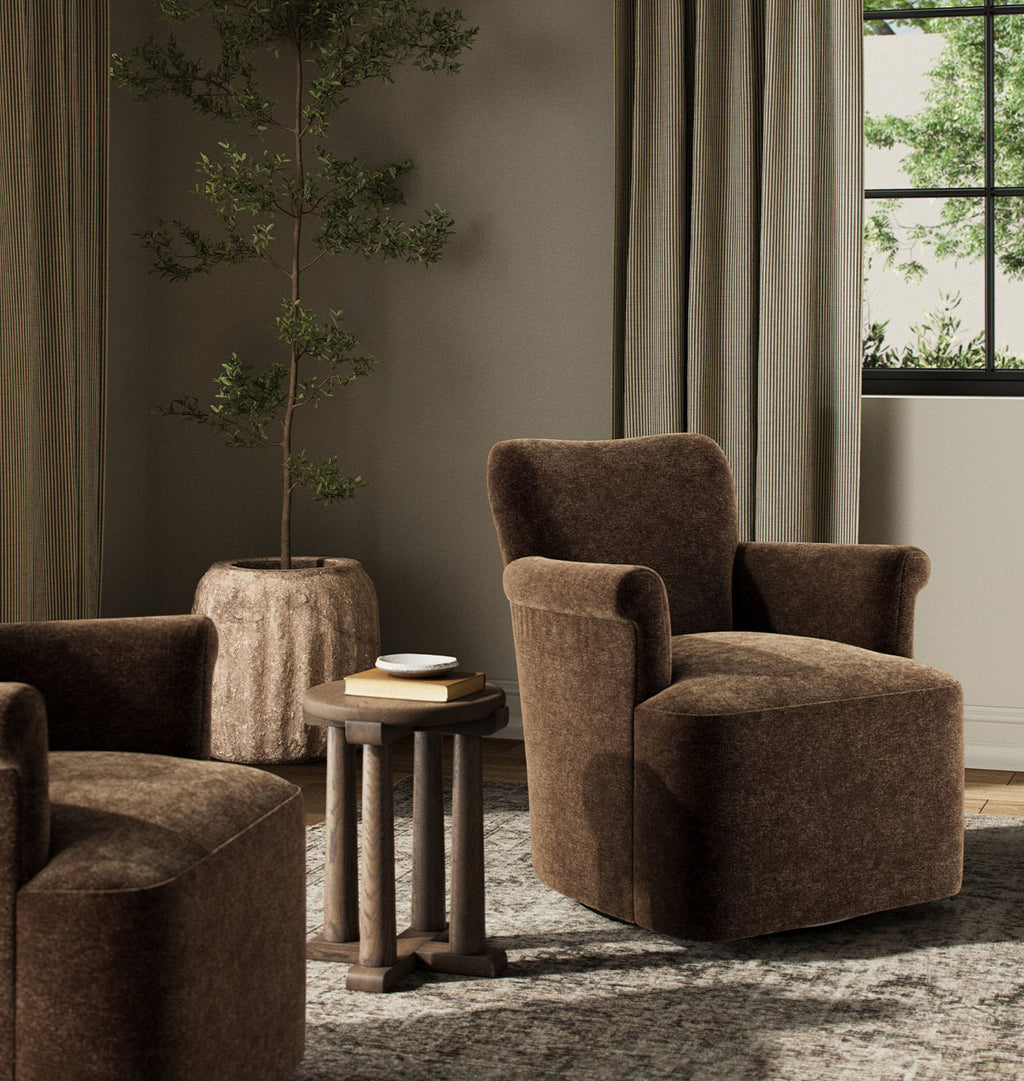 A brown upholstered armchair in a styled living room setting, alongside a matching side table and another chair, with curtains, a potted tree, and a window in the background.