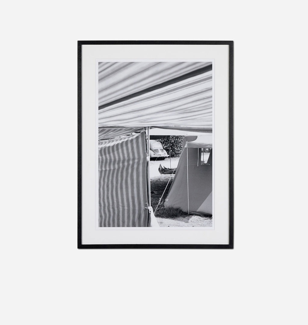 A framed black and white photographic print featuring a striped awning and a tent, presented in a black frame against a light gray background.