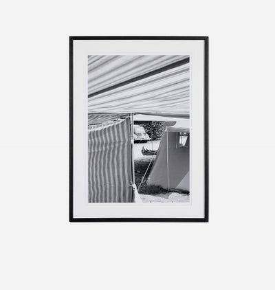 A framed black and white photographic print featuring a striped awning and a tent, presented in a black frame against a light gray background.