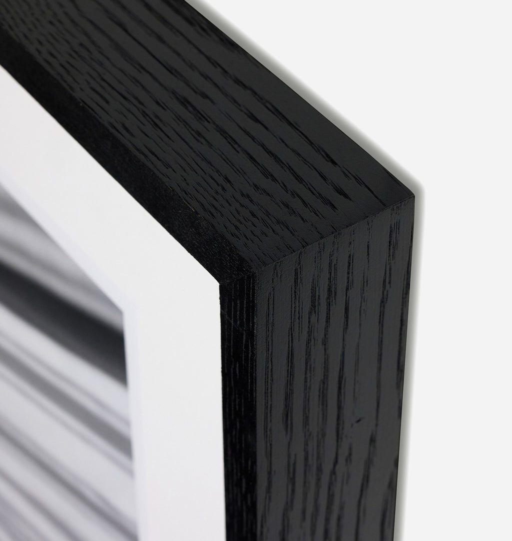 Close-up of a black wood frame corner with a white mat and a black-and-white striped artwork visible inside, on a light gray background.