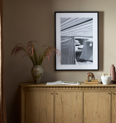 A framed black and white architectural photograph hanging on a warm taupe wall above a light wood cabinet, styled with a vase of ornamental grass, an open book, and small decorative objects.