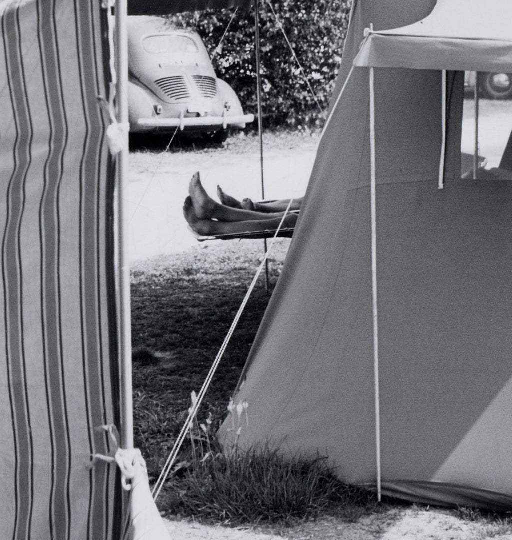 Black and white photograph of a camping scene with a striped beach chair on the left, a large tent on the right, and a person lying on a hammock strung between the tent and the chair. A vintage car is parked in the background near some foliage.